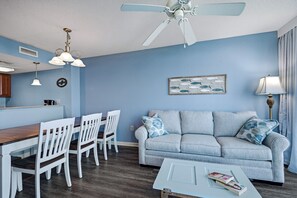A living room with a blue wall, white furniture, a ceiling fan, and a coffee-themed wall decoration. - Dunes Village 452 - Updated Oceanfront 2BR (Myrtle Beach)