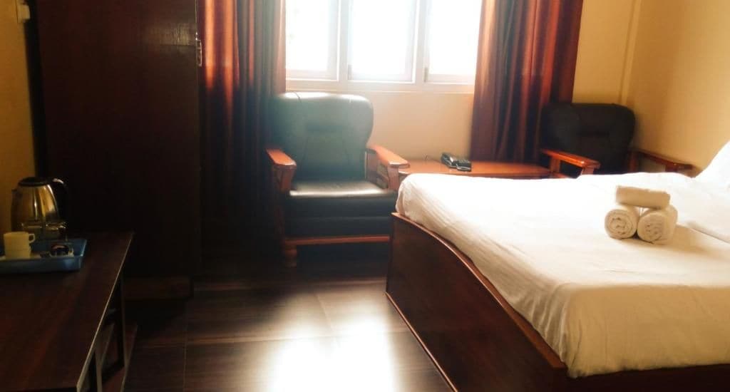 Una habitación de hotel con una cama, un escritorio con hervidor, una silla y un ventanal con cortinas.
