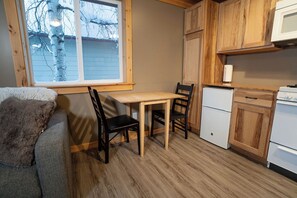 A compact kitchen with wooden cabinets, a small refrigerator, and a dining table with chairs. - Cozy cabin in the heart of Talkeetna - perfect for any getaway (Talkeetna)