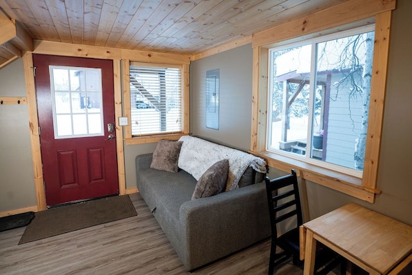 Ein kleiner Raum mit roter Tür, grauem Sofa mit Decke, einem Holztisch und einem Fenster mit Jalousien.