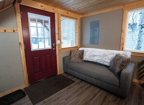 A room with a red door, a grey sofa with two pillows, and a window with blinds. - Cozy cabin in the heart of Talkeetna - perfect for any getaway (Talkeetna)