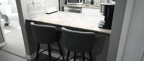 A kitchen with a granite countertop, two bar stools, a microwave, and a coffee maker.