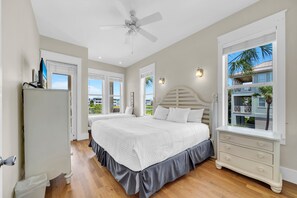 Ein Schlafzimmer mit Bett, Kommode, Deckenventilator und Fenster mit Blick auf Gebäude und Palmen.