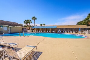 Una piscina con sillas de descanso y una zona cubierta, rodeada de palmeras y una valla de madera.