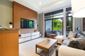 A modern living room with a flat-screen TV mounted on a wooden wall, a sofa, a coffee table, and a large window offering a view of greenery. - 4BR Ski Chalet, 2472 Sq Ft, w/ Pvt HotTub & Garage (Whistler)