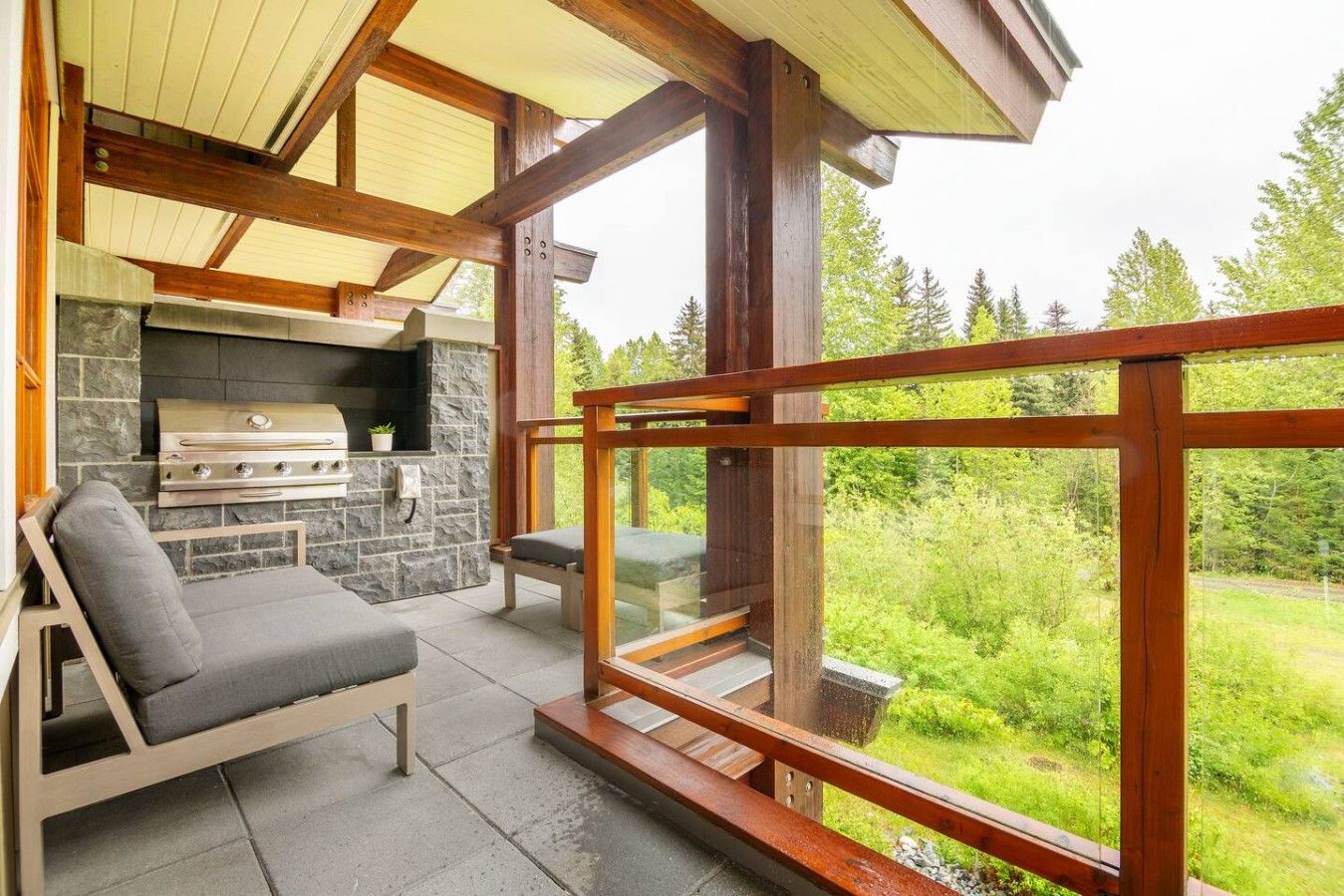 A covered outdoor seating area with a stone fireplace, a grill, and a view of a forested landscape.
