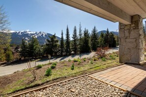 A balcony with a stone pillar, a paved area with gravel, and a view of a road with trees and snow-capped mountains in the distance. - Stunning 4BR Chalet w/ Mountain Views, Pvt Garage (Whistler)