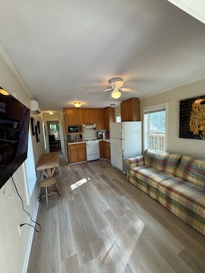 A compact kitchen with wooden cabinets, a microwave, stove, and refrigerator. There's a plaid sofa and a dining area with a stool. - Bayou Bengal Bungalow Fire Pit-Grill-Front & Back Porch Near LSU & Restaurants (Baton Rouge)