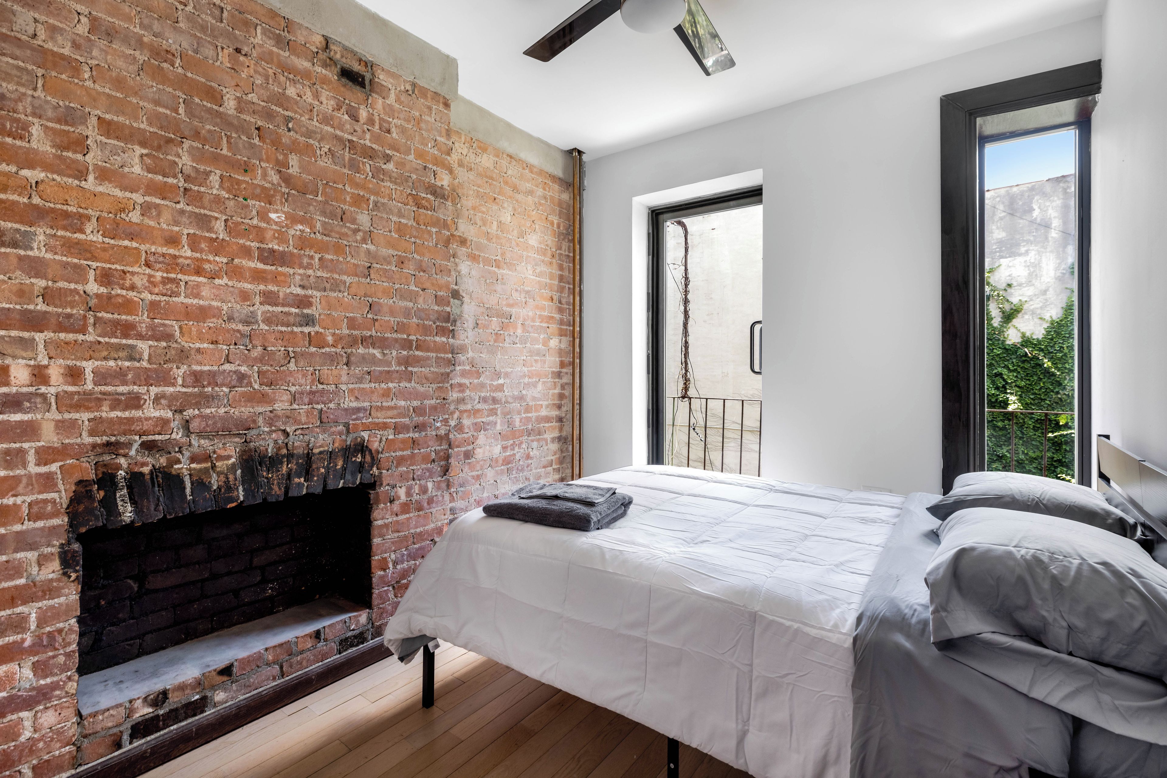 Un dormitorio con chimenea de ladrillo, una cama con ropa blanca y vistas a zonas verdes a través de una ventana.