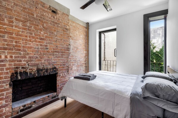 A bedroom with a brick fireplace, a bed with white bedding, and a view of greenery through a window.