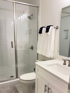 A modern bathroom with a glass shower enclosure, a toilet, a sink with a mirror, and two towels on a towel rack. - 2-Bedroom Condo in Auburn-Walk to Stadium and Downtown (Auburn)