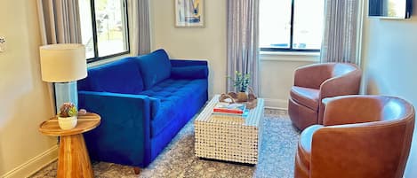 Una sala de estar con un sofá azul, un sillón de cuero marrón, una mesa redonda con libros y una lámpara de pie.