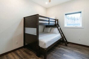 A bunk bed with a ladder, a window with a view of trees, and a wooden floor. - The Alpine Den (Revelstoke)