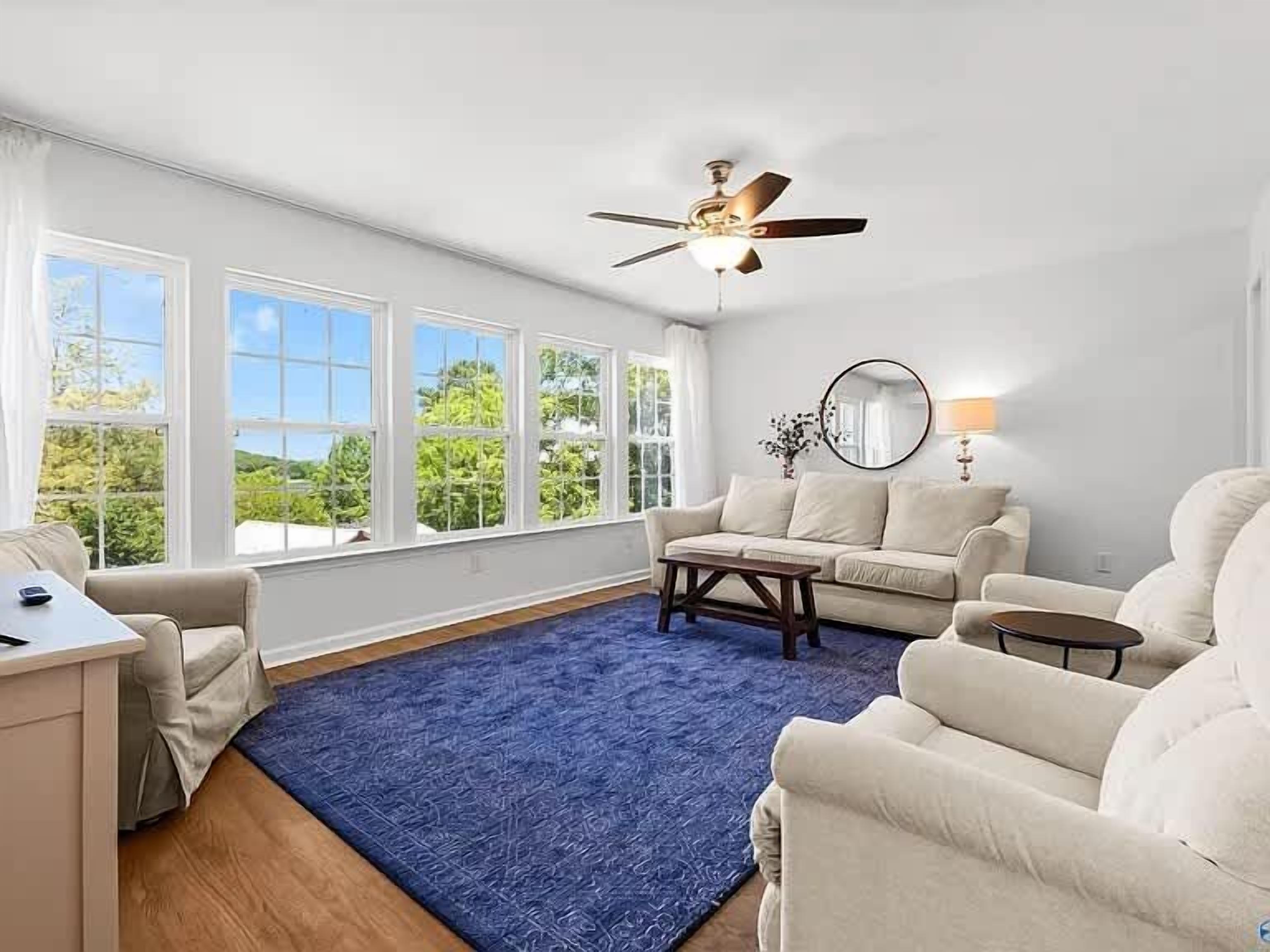 A spacious living room with a blue rug, a ceiling fan, a sofa, a coffee table, and a view of the outdoors through large windows.
