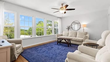 A spacious living room with a blue rug, a ceiling fan, a sofa, a coffee table, and a view of the outdoors through large windows.