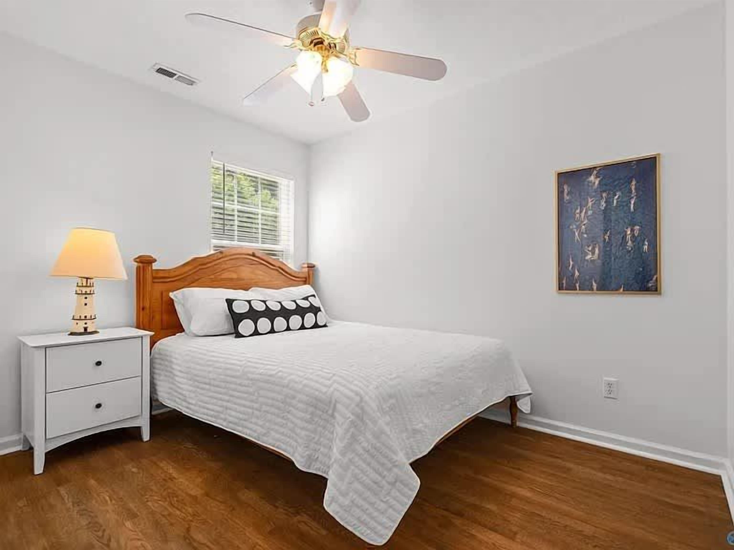 A bedroom with a bed, nightstand, lamp, ceiling fan, window, and artwork on the wall.