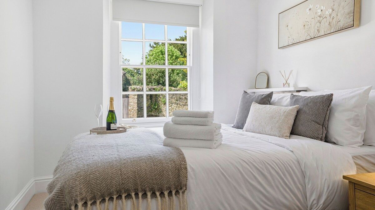 A bedroom with a large window, a bed with white linens, a bottle of champagne, and a framed picture on the wall.