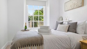 Quarto com uma janela grande, cama com lençóis brancos, uma garrafa de champanhe e um quadro na parede.