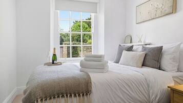 A bedroom with a large window, a bed with white linens, a bottle of champagne, and a framed picture on the wall.