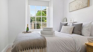 Un dormitorio con un ventanal grande, una cama con ropa blanca, una botella de champán y un cuadro en la pared.