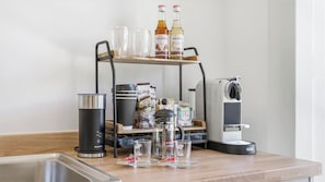 Una encimera de cocina con cafetera, hervidor, taza y estante con botellas y vasos.