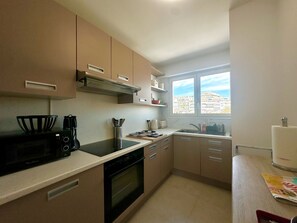 A modern kitchen with a microwave, stove, and sink. There is a window providing a view of buildings outside. - Charmant 3P vue mer balcon panoramique, Clim (Cannes)