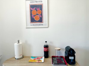 A wall with a framed artwork titled 'LES FRUITS LA MANDARINE' and a table with a tissue roll, a bottle of Coca-Cola, a glass jar, a magazine, and a coffee machine. - Charmant 3P vue mer balcon panoramique, Clim (Cannes)