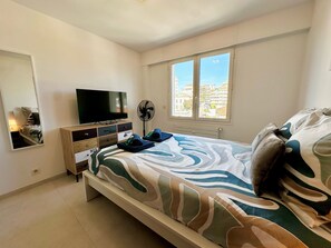 A bedroom with a bed, a TV, a fan, and a window with a city view. - Charmant 3P vue mer balcon panoramique, Clim (Cannes)