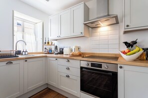 A modern kitchen with white cabinets, a wooden countertop, a built-in oven, and a window with curtains. - The Cottage at Old Coombe House (Tetbury)