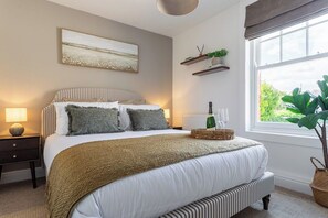 A bedroom with a bed, bedside tables, a window with a view, and a wall-mounted shelf with plants. - New! Stylish King Bed Apartment | Central Lechlade (Lechlade)