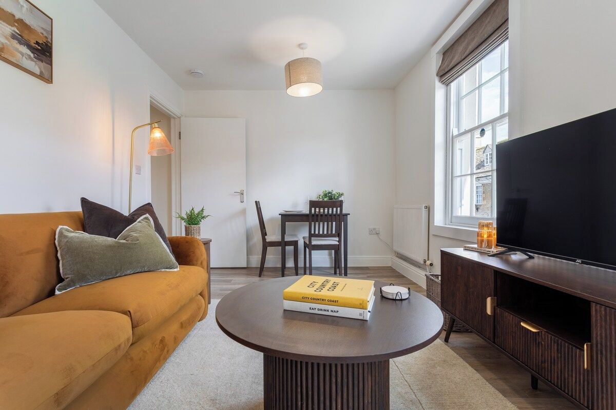A living room with a sofa, a coffee table with books, a TV, and a dining area with chairs and a table.