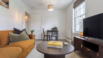 A living room with a sofa, a coffee table with books, a TV, and a dining area with chairs and a table.