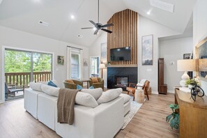 A modern living room with a fireplace, a large TV mounted on a wooden wall, and a ceiling fan. - Trailfront Ride & Reel - Upscale Retreat 4bd/2.5ba Sleeps 10 - HOT TUB! (Bella Vista)