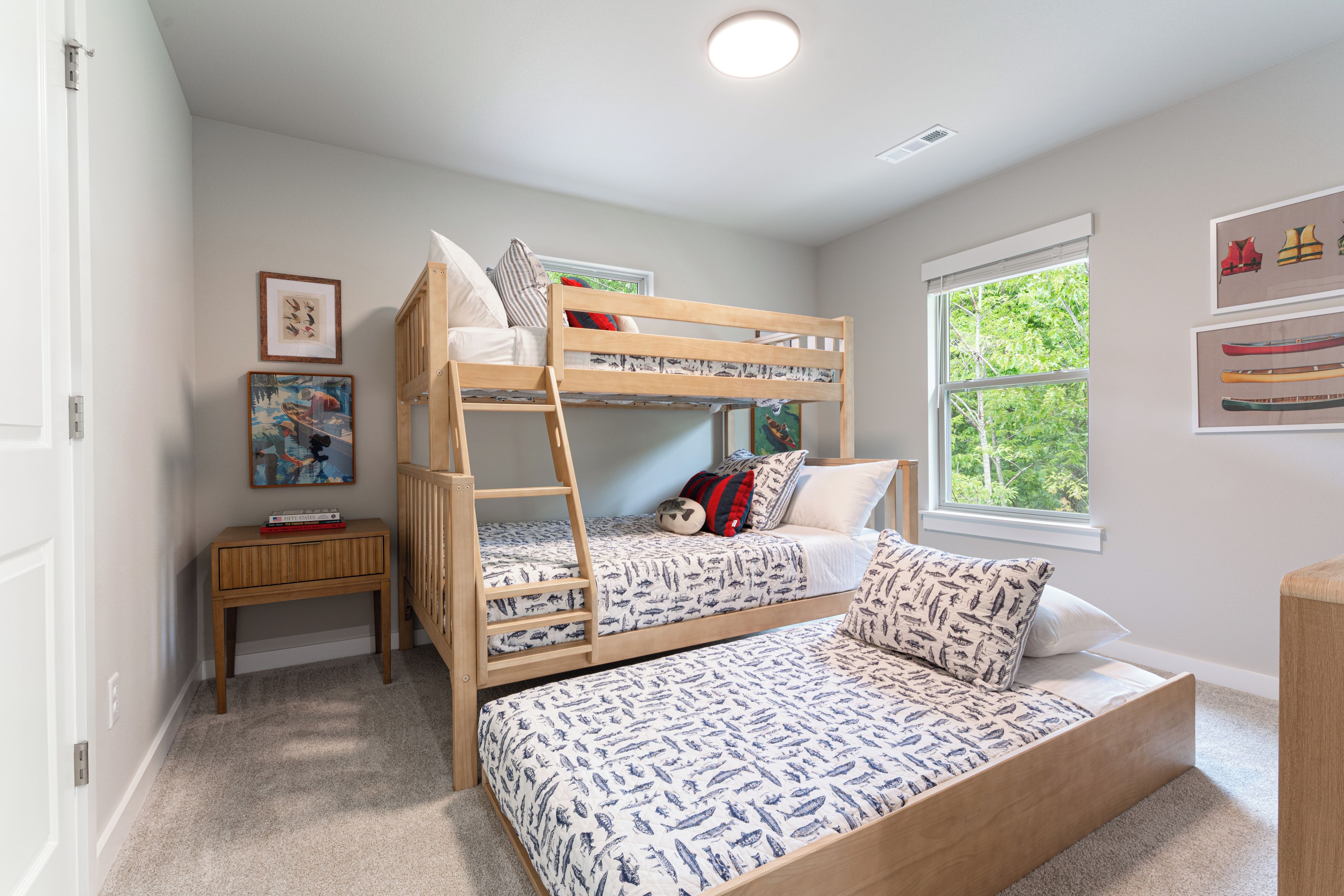 Habitación infantil con literas, una mesita de noche, una ventana con vistas a árboles y cuadros en la pared.