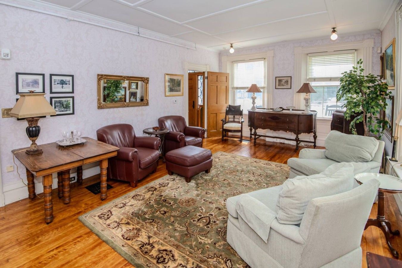 Ein Wohnzimmer mit einem Holztisch, Ledersesseln, einem gemusterten Teppich, einer Couch, einem Klavier und gerahmten Kunstwerken an den Wänden.