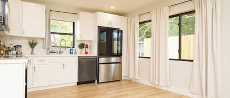 A modern kitchen with white cabinets, stainless steel appliances, and large windows with curtains.