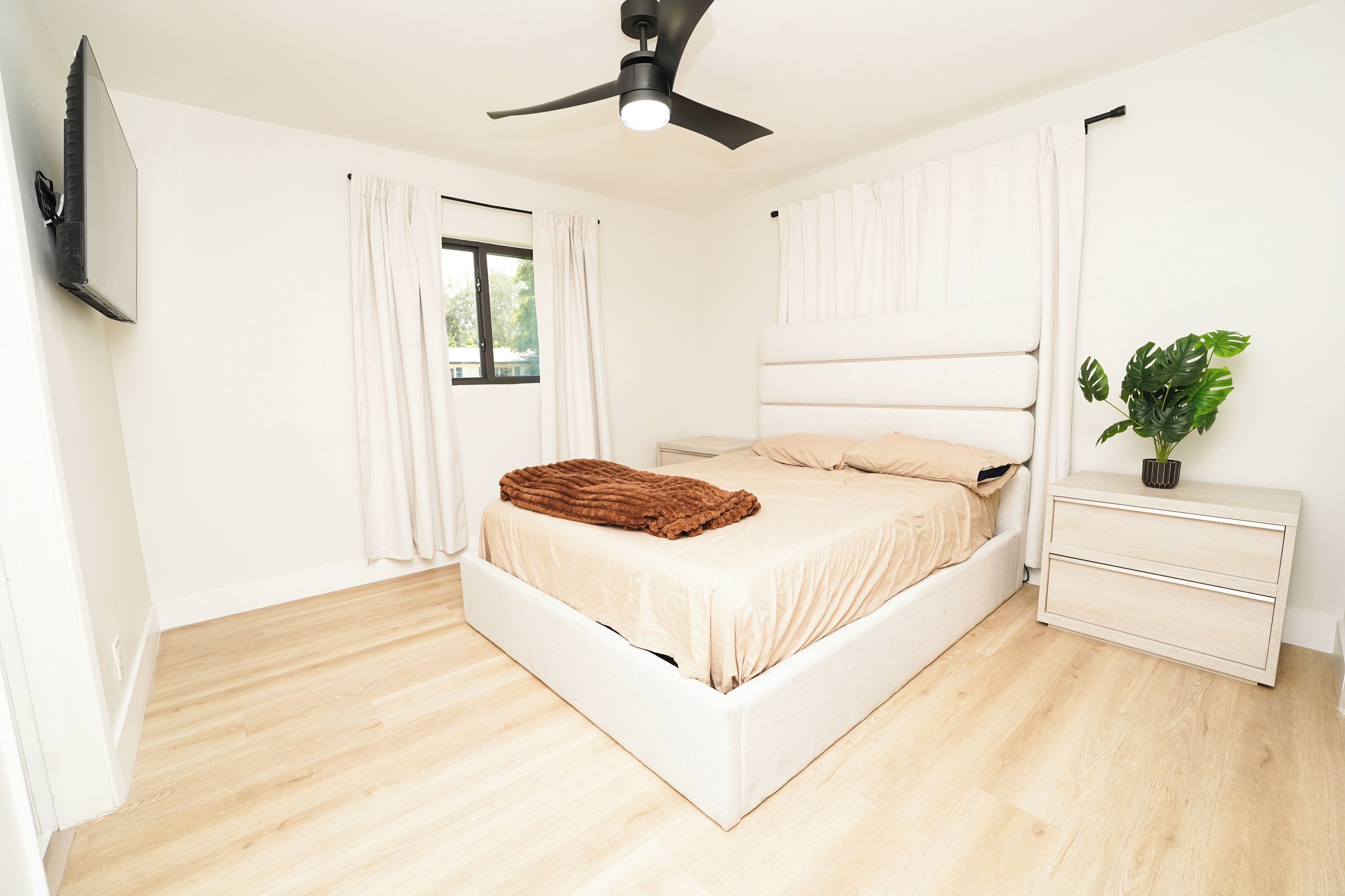 Un dormitorio con cama, mesita de noche, ventilador de techo y ventana con cortinas.