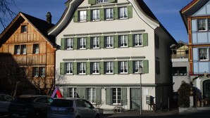 A multi-story building with a steep gable roof, multiple windows with shutters, and a visible entrance with a door. - Wohnung mit Sicht auf den Landsgemeindeplatz. (Appenzell)