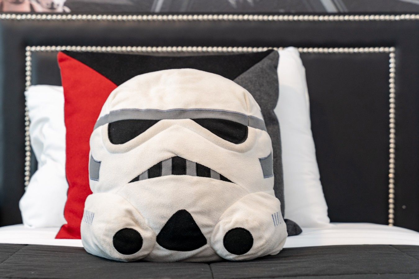 A Stormtrooper pillow on a bed with a black headboard and a red pillow.