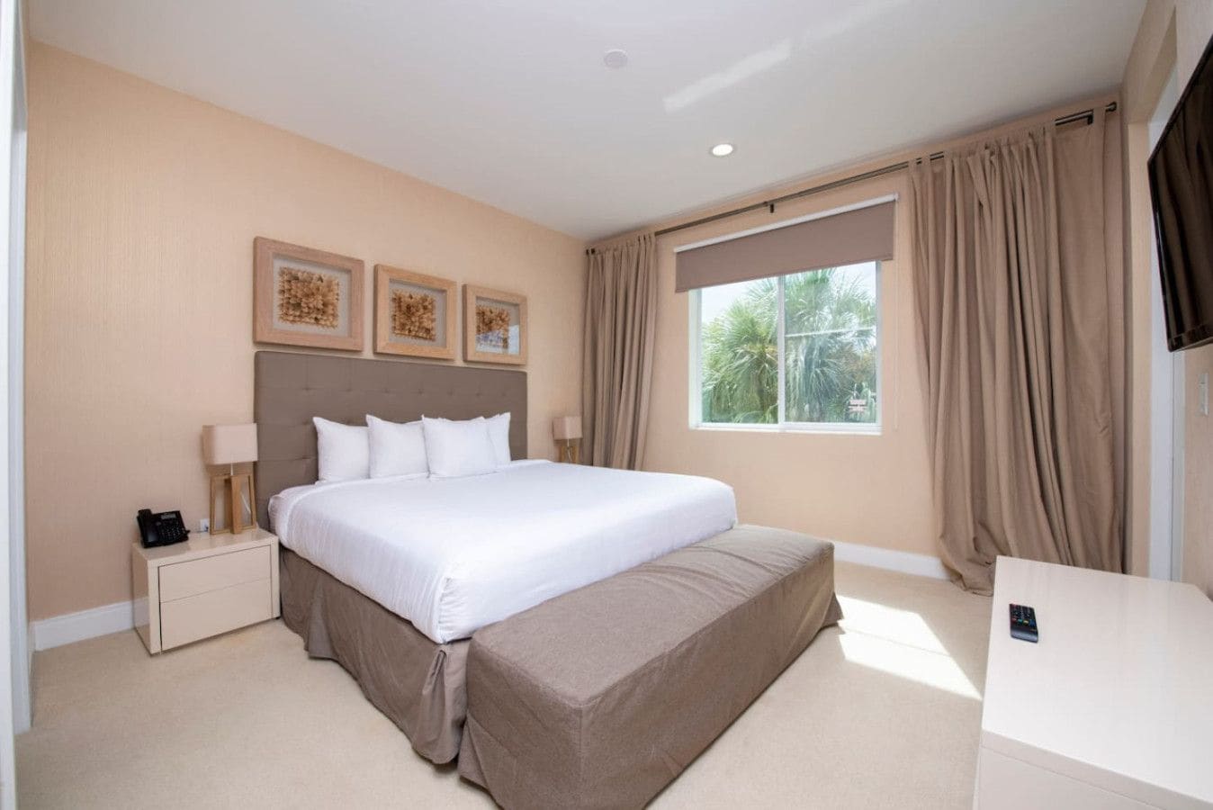 Quarto de hotel com uma cama grande, mesa de cabeceira com abajur, televisão e janela com cortinas.