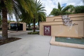 Entrada de un complejo turístico con palmeras, un elemento de agua y un cartel que dice 'Magic Village Resort'.