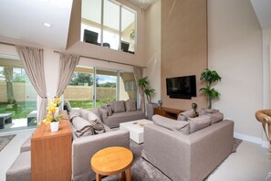 Ein modernes Wohnzimmer mit hoher Decke, großen Fenstern und einem an der Wand befestigten Flachbildfernseher.