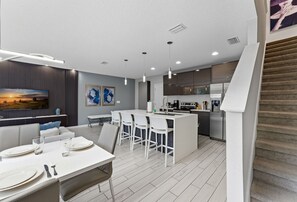 Una cocina moderna con armarios blancos, una isla central con taburetes y un comedor con una mesa blanca dispuesta para cuatro.