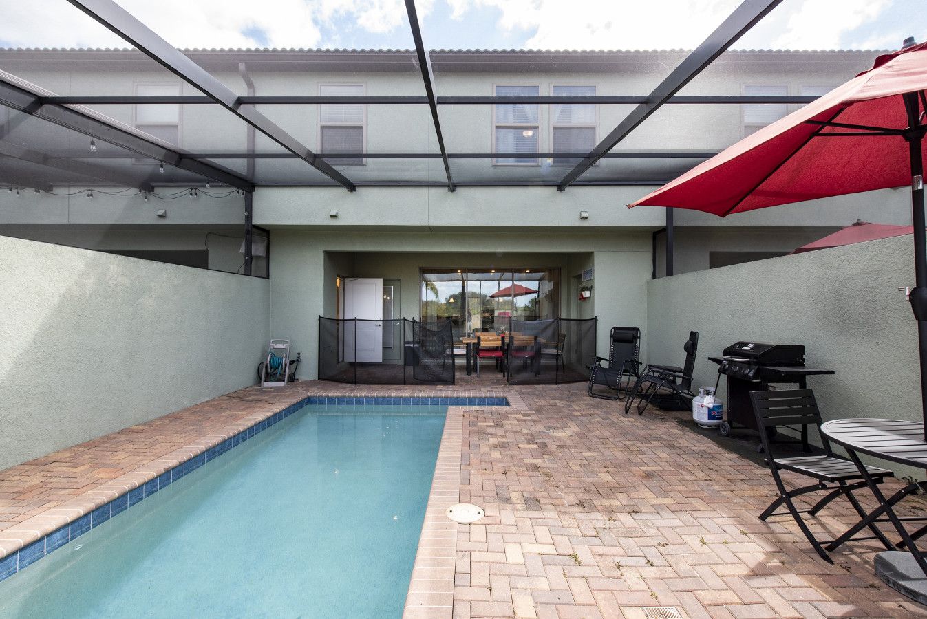 Une piscine dont le bord est revêtu de carreaux, entourée d’une terrasse en briques. On y trouve un espace de détente couvert avec un parasol rouge, un barbecue et du mobilier d’extérieur.