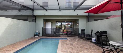 A swimming pool with a tiled edge, surrounded by a brick patio. There is a covered seating area with a red umbrella, a grill, and outdoor furniture.