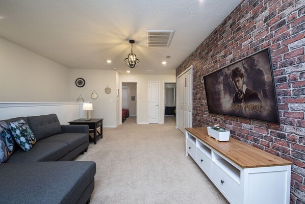 A living room with a grey sofa, a wooden coffee table, a TV mounted on a brick wall, and a hallway leading to another room. - 5BD Family Home with Pool BBQ and Themed Rooms (Davenport)