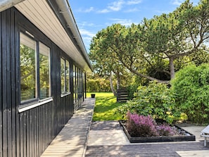 A modern house with a wooden deck, large windows, and a well-maintained garden with various plants and trees. - 6 Person Holiday Home in Juelsminde (Juelsminde)