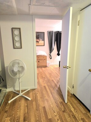 Een kamer met een houten vloer, een staande ventilator, een bed met een wit kussen en een deur naar een andere kamer.