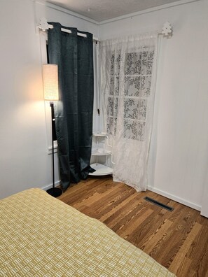 Ein Schlafzimmer mit einem Bett, einem Nachttisch, einer Lampe, einem Fenster mit Vorhängen und einer Heizung.