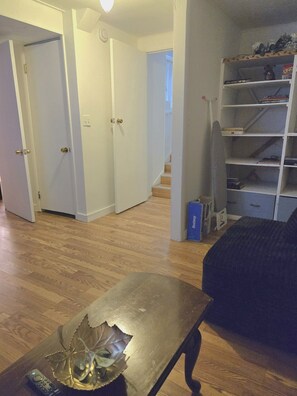 Een kamer met een houten vloer, een zwarte bank, een bijzettafeltje met een decoratief object, een boekenkast en een trap die naar een andere kamer leidt.
